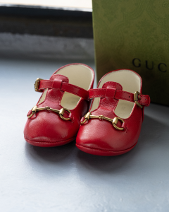 Red Gucci baby shoes with gold accents, placed beside a green Gucci box on a soft-lit surface.