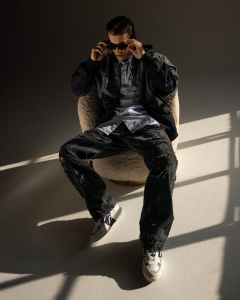 A man in a dark outfit with sunglasses sitting on a chair, light falling dramatically.